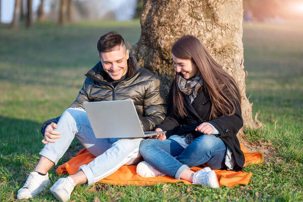 Красивая пара в повседневной одежде веселится с ноутбуком на улице. Сидя у подножия дерева, вы наслаждаетесь природой в солнечный весенний день. Концепция удаленной командной работы.