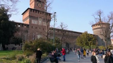Milan, İtalya, Mart 2022. Sforza kalesinin eğik görüntüleri. İnsanlar güneşli günün tadını çıkarıyorlar, saatli kırmızı tuğlalı cephe ve çan kulesi kanıt..