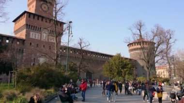 Milan, İtalya, Mart 2022. Sforza kalesinin eğik görüntüleri. İnsanlar güneşli günün tadını çıkarıyorlar, saatli kırmızı tuğlalı cephe ve çan kulesi kanıt..
