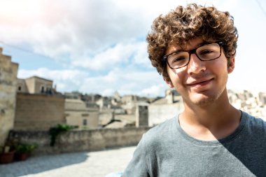 Matera, Basilicata, İtalya. Ağustos 2021. Genç bir turist kameraya bakar ve gülümser. Arkasındaki tarihi merkez bulanıklaşır. Tatil ve tasasız konsept. Boşluğu kopyala.