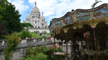 Paris, France, June 2022. Graceful footage with a nice view of the Sacre Coeur basilica, framed by the canopy of trees on the left and on the right the typical Parisian vinatge carousel as it rotates.