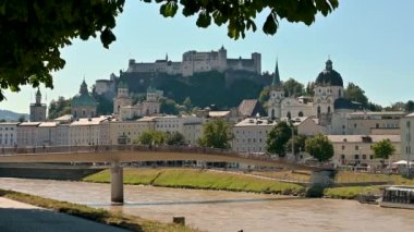 Salzburg, Avusturya, Ağustos 2022. Ağaçlar ve nehir tarafından çerçevelenmiş eski bir kasabanın görüntüsü ile şaşırtıcı gündüz görüntüleri. Güzel bir yaz günü.