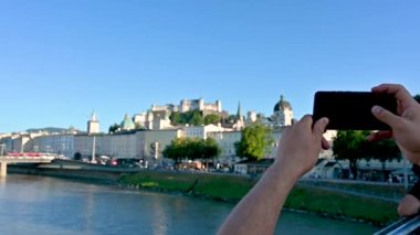 Salzburg, Avusturya, Ağustos 2022. Ön planda ön planda fotoğraf çekmek için elinde cep telefonuyla gündüz çekilen yavaş çekim görüntüleri. Arka planda, kalenin egemen olduğu eski kasaba.