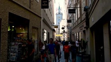 Salzburg, Avusturya, Ağustos 2022. Merkezin sokaklarını araştıran POV görüntüleri, alışveriş caddesi boyunca tarihi işaretlerle insanlar arasında yürüyoruz. Arka planda bayraklı belediye kulesi var..