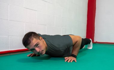 Şınav çeken ve kameraya bakan kararlı genç bir adam. O, gri bir gömlek ve siyah bir tulum giydi. Boşluğu kopyala