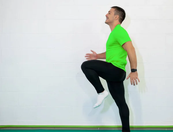 Work out. Young caucasian male athlete sprinter running, exercising indoors, jogging in training room, side view. Green shirt and black jumpsuit. Copy space