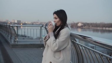 Ceket ve şapka giyen neşeli, genç bir kızın şehir caddesinde durup kahve içişini yakından çek.