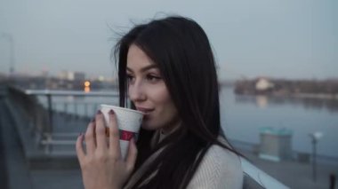 Güzel bir kız sıcak bir ceketle sokakta dikiliyor. Elinde kahve fincanı var ve başka tarafa bakıyor.