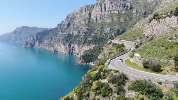 Sparatoria aerea di vista perfetta di strada tra riva rocciosa verso montagne. Auto bianca lungo la strada della Costiera Amalfitana, Italia. Video 4K. — Video Stock