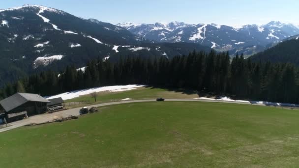 Malerische Luftpanorama-Landschaft in den Alpen mit saftig grünen Almen und schneebedeckten Berggipfeln im Hintergrund. Ein schwarzes Auto fährt auf ein Haus auf einem Berg zu. 4K-Video. — Stockvideo