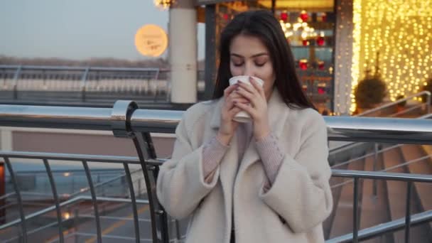 Outside shot of satisfied attractive female model with healthy skin, brown hair, holds takeaway coffee, enjoys good drink, dressed in jacket, waits for someone, has coffee break, looks at camera. — Stock Video