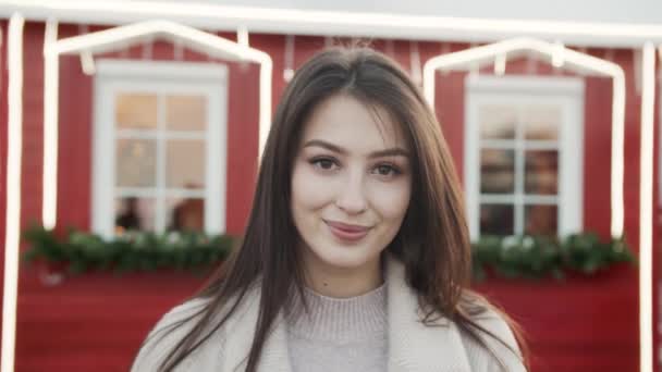 Schöne lächelnde Brünette steht auf der Straße in der Nähe des roten Hauses mit Laternen für Weihnachten oder Neujahr dekoriert — Stockvideo