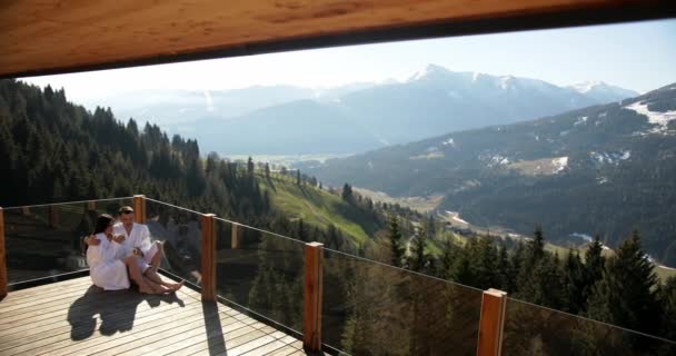 Hermosa pareja feliz sentada en el balcón de su cabaña de montaña de vacaciones mientras bebe café y disfruta de la vista a la montaña — Vídeos de Stock