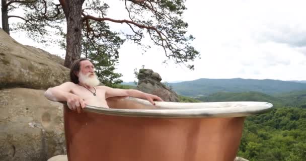 Ein erwachsener Mann Weihnachtsmann sitzt in einer Badewanne in den Bergen an der frischen Luft. — Stockvideo