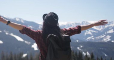 Arkadan bakıldığında, kaygısız esmer bir kadının, temiz havanın serin esintisinden zevk alırken kolları geniş, dağların ve ağaçların güzel manzarasını izleyen bir şapka taktığı görülüyor..