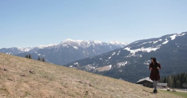 Wisatawan berambut cokelat perempuan mengenakan topi berjalan di padang rumput sambil mengamati pemandangan di sekitarnya dengan latar belakang pegunungan yang indah. — Stok Video