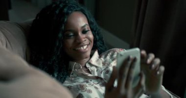 Kanepede uzanmış kıvırcık saçlı, akıllı telefon cihazıyla mesajlaşırken ölçü aldıktan sonra kahkahalarla gülen genç ve çekici bir Afro-Amerikan kadının portresi. Bir yanıtın düşüncesi.