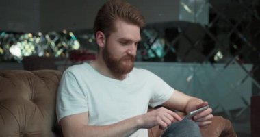 Sakallı beyaz adamın akıllı telefonundan süzülüşünü, evdeki kanepede otururken bir görüntüye yakınlaşmasını yakından izle..