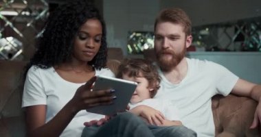 Mutlu bir melez çiftin kanepede yan yana oturduğu yakın plan. Yakışıklı oğulları gülüyor, gülümsüyor ve akıllı telefondan video seyrediyor..