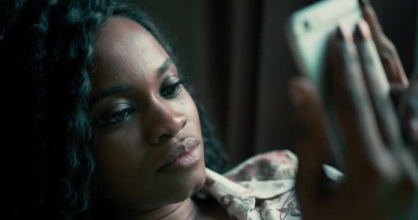 若い魅力的なアフリカ系アメリカ人女性の超クローズアップポートレートで、手にスマートフォンのデバイスにテキストメッセージを送りながらメッセージを受け取った後、ソファの上で笑って横になって巻き毛を持つ。Aの事 — ストック動画