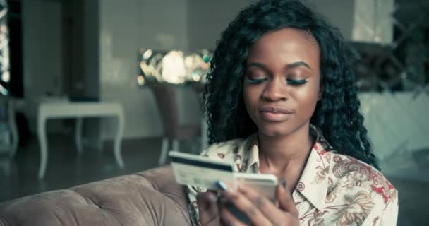Close up retrato de uma bela mulher afro-americana com cabelo encaracolado entrando seus detalhes de cartão de crédito em seu smartphone enquanto faz compras on-line. Interior. — Vídeo de Stock