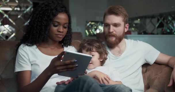 Vista frontal de cerca de una feliz pareja de carreras mixtas sentados en un sofá junto con su lindo hijo riendo, sonriendo y viendo un video en un teléfono inteligente. — Vídeo de stock