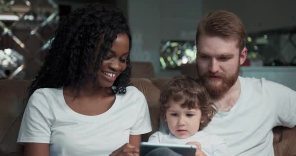 Close-up widok z przodu szczęśliwej mieszanej pary wyścigowej siedzi na kanapie razem z ich cute syn śmieje się, uśmiecha i gra w grę na smartfonie. — Wideo stockowe