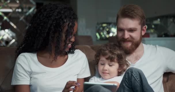 Vista frontal de cerca de una feliz pareja de razas mixtas sentados en un sofá junto con su lindo hijo riendo, sonriendo y jugando un juego en un teléfono inteligente. — Vídeos de Stock