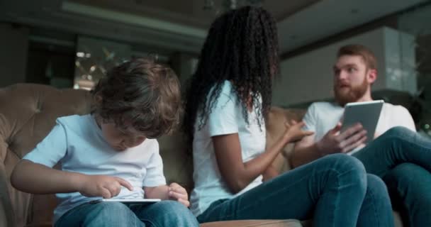 Triste niño sentado jugando juegos en un teléfono mientras sus padres pelean en casa en la sala de estar. — Vídeos de Stock