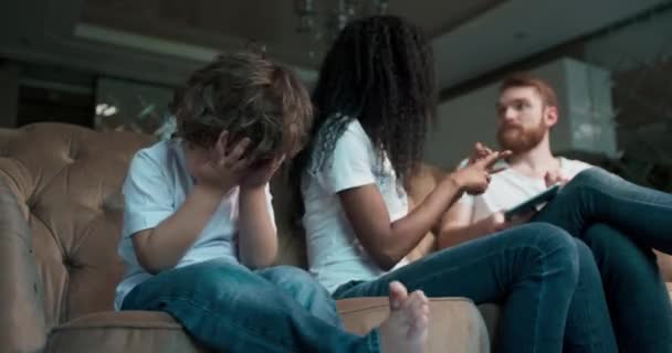 Sorglig och irriterad liten pojke sitter bredvid sina föräldrar när de slåss hemma i vardagsrummet. — Stockvideo