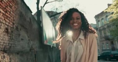 Kıvırcık saçlı, güzel, mutlu, Afro-Amerikan bir bayanın kamera karşısına geçişi. Güneşli bir günde dışarıda eğlenmek..
