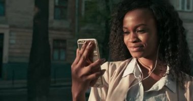 Açık havada dikilirken akıllı telefonuyla görüntülü sohbet ederken gülümseyen güzel bir Afro-Amerikalı kadının yakın görüntüsü..