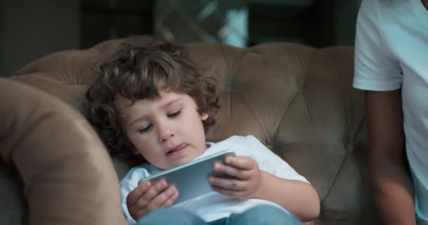 Lindo niño jugando juegos en un teléfono inteligente y su madre afroamericana se inclinan para ayudarlo. — Vídeo de stock