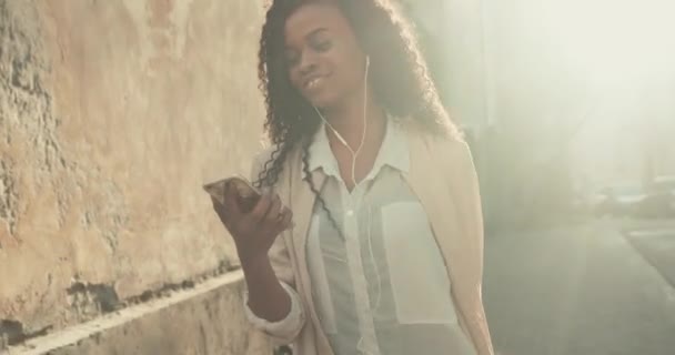 Вид Баклита на веселую афроамериканку, идущую по улице с мини-чемоданом, слушая музыку. — стоковое видео