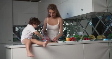 Mutfak tezgahında oturmuş domateslerle oynayan küçük bir çocuğun yakın plan görüntüsü. Annesi akşam yemeği için sebzeleri kesiyor..