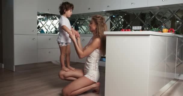 台所でスクワットをしながら、幼い息子を足で跳ねている母親の映像。母親と息子の絆の時間. — ストック動画