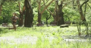 Odaklanmış sarışın bir jimnastik salonunun, ağaçlarla çevrili çimenli bir alanda koşuşunun yavaş çekim görüntüleri. Kardiyo çalışma rutini.