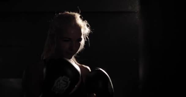 Yakın çekim arka ışıklandırması, antrenman sırasında boks çantasına yumruk atan kararlı bayan boksör.. — Stok video