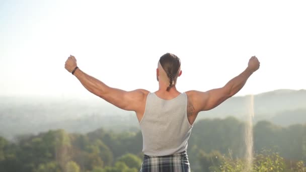 Rear view. a strong man with large muscles in his hands spreads the hands to the sides and from his fists pours sand. the wind blows the sand. the background of the big forest below — Stock Video
