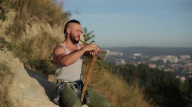 Sakallı bir viking gibi çekici bir adam elinde baltayla bir kayanın üzerinde oturuyor. Adam gökyüzüne bakıyor. yerleşim arka planda görünür..