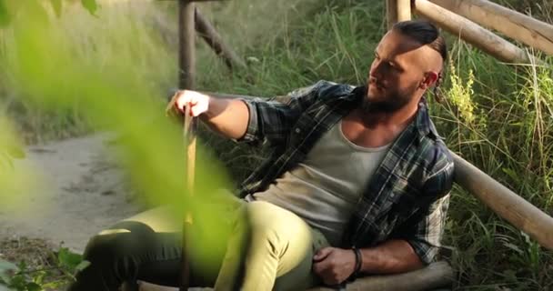 Atrakcyjny mężczyzna w czystych ubraniach siedzi na ścieżce opierając się o drewnianą tratwę z siekierą w rękach — Wideo stockowe