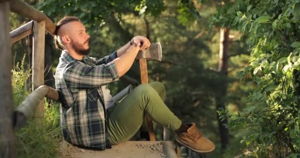Den unge mand er ked af det. en mand sidder med ryggen mod en træbro og hovedet først på en økse. ung mand med en Viking-lignende udseende – Stock-video