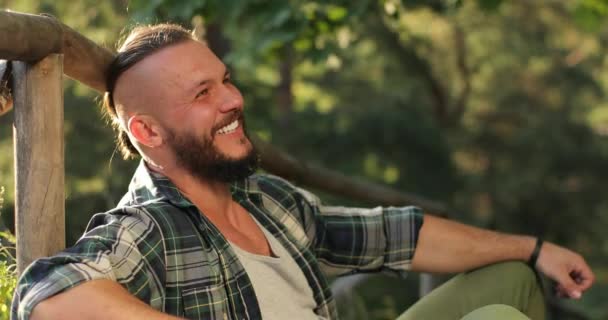 Ένας ελκυστικός άντρας με όμορφο χτένισμα κάθεται στη φύση ακουμπισμένος σε μια ξύλινη σχεδία και γελώντας — Αρχείο Βίντεο