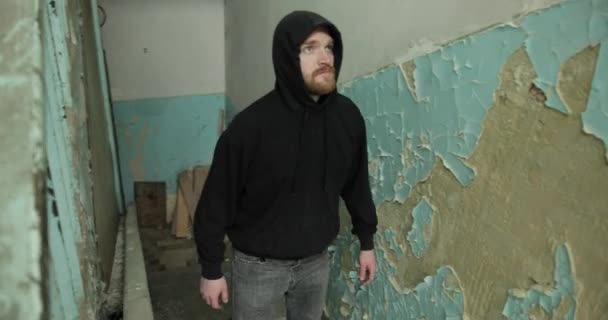 Человек с бородой в черном свете и серых джинсах поднимается по лестнице в полуразрушенный дом. молодой человек оглядывается на разрушенные стены — стоковое видео