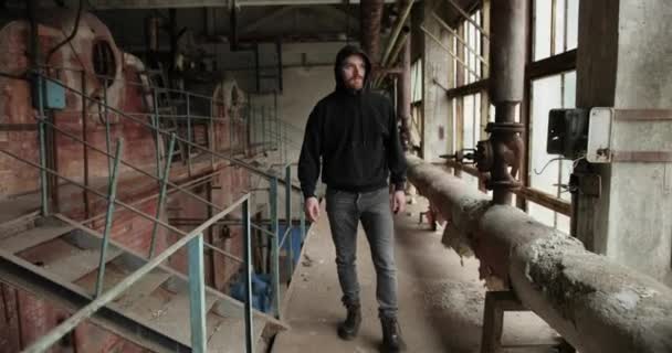 Ein junger bärtiger Mann mit Kapuze geht in die zerstörte Fabrik. um schäbige Wände, gerissene Drähte, rostige Rohre und Geländer. — Stockvideo