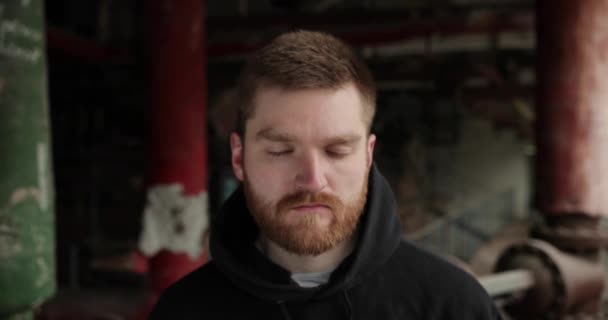 Nära porträtt av en man med rött skägg. Man står med slutna ögon i en övergiven fabrik. allvarliga ansiktsuttryck — Stockvideo