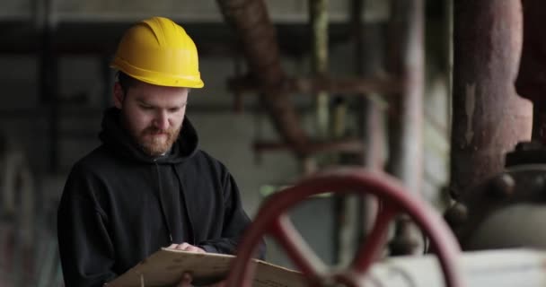 Travailleur en casque jaune examine les documents. homme se tient à une usine près d'un robinet d'eau. ingénieur examine les documents et sourit — Video