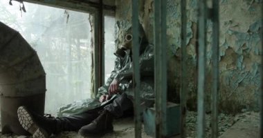 Gaz maskeli ve tulumlu bir adam pencerenin yanındaki yarı çökmüş bir odada duvara yaslanmış oturuyor.