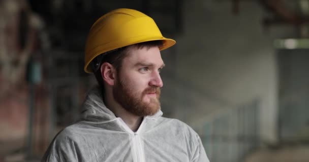 Profile of a man in a white coat in a yellow helmet stands indoors and smiles — Stock Video