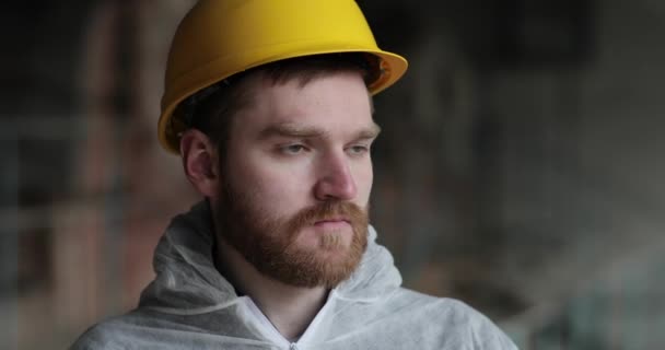 Skäggig ung man i vit rock och gul hjälm tittar in i kameran och ler. fabriksingenjör. befälhavaren i kistan — Stockvideo